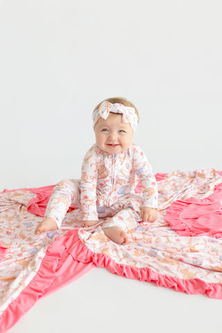 Infant sitting on blanket with boots and flower print pattern, ruffled pink trim along edges.