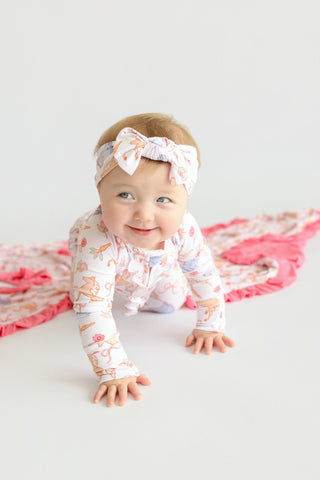 Infant wearing white long-sleeve romper with pink and orange floral print, matching headband with bow, crawling on white surf