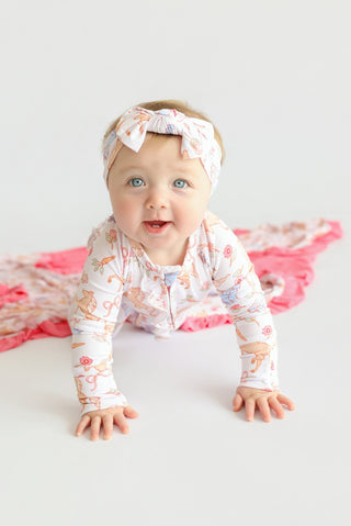 Infant wearing white romper with floral print, matching headband, crawling on white surface with pink ruffled fabric.