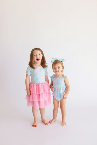 Toddler wearing blue and pink bubble romper with matching hair bow standing next to older child in layered pink tulle skirt.