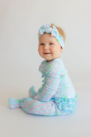 Infant wearing light blue romper with multicolored floral and geometric print, matching headwrap, and ruffled leg cuffs.