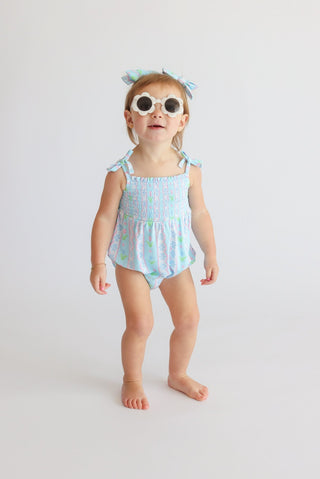 Toddler wearing light blue romper with tulip print and smocked bodice, paired with white round sunglasses and coordinating ha