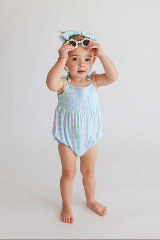 Toddler wearing light blue bubble romper with tulip print and smocked bodice, paired with matching hair bow and sunglasses.