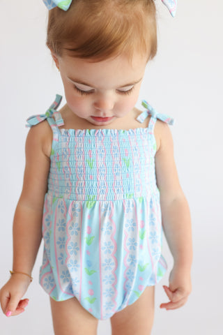 Toddler wearing light blue bubble romper with smocked chest, tie shoulder straps, and multicolor floral tulip print throughou