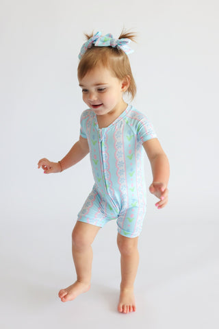 Toddler wearing short-sleeved romper with tulip and geometric pattern in blue and pink with matching bow headband.