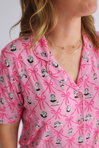 Pink short-sleeve top with ribbon bow and figure print pattern, worn with layered gold necklaces.