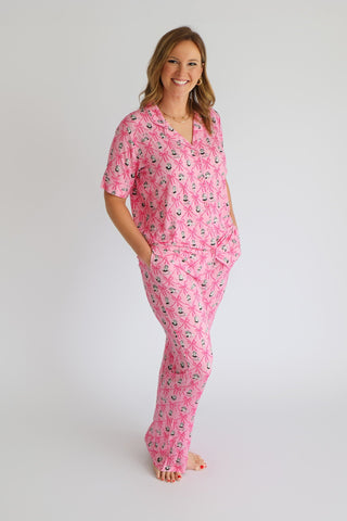 Woman wearing pink pajama set with ribbon and crown print, short-sleeved top and flare pants.