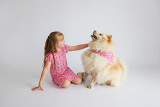 Pink dog bandana with small pattern, tied around tan and white dog's neck next to child in matching pink outfit.
