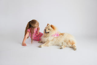 Cream and tan dog wearing pink bandana with ribbon pattern, posed with child in matching pink outfit.