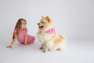 Cream and tan dog wearing pink bandana with ribbon pattern, sitting next to young child in matching pink outfit.