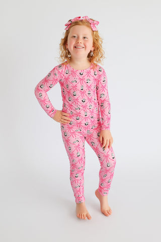 Girl wearing pink long-sleeve pajama set with ribbon and ballerina print, matching pink hair bows, and pearl earrings.