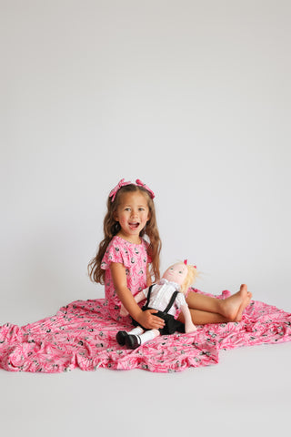 Pink ruffle blanket with small scattered pattern, displayed with child and doll on gray background.