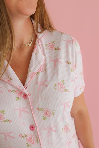 Woman wearing white ribbed button-up shirt with pink roses and bows print, pink trim, and matching necklace against pink back