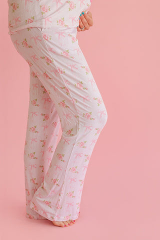 Woman wearing cream ribbed pajama pants with pink floral and bow print, shown from waist down against pink background.