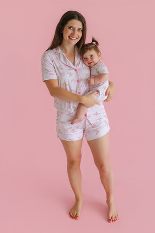 Woman and toddler wearing matching pink floral short pajama sets with button-up tops against a pink background.