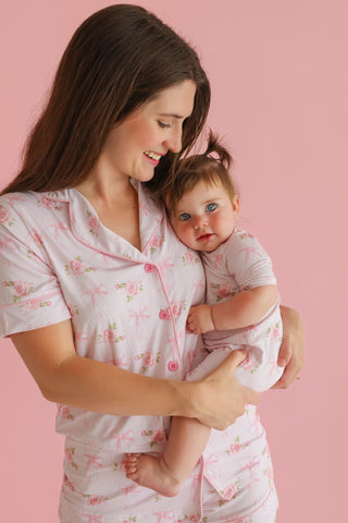 Woman and infant wearing matching pink floral short sets with button-front tops against pink background.
