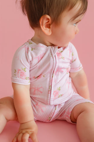 Toddler wearing pink short-sleeve romper with floral rose print and bow details on pink background.