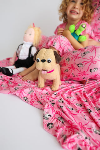Pink ruffle blanket with floral and dog print pattern displayed with child and plush toys.