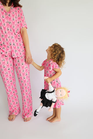 Woman and child wearing matching pink pajama sets with ribbon and carousel horse print, child holding doll.