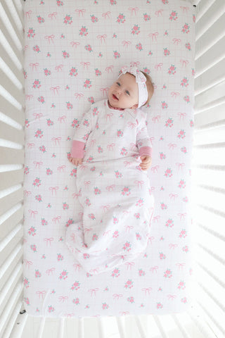 Infant in white sleep sack with pink rose and bow pattern, lying in white crib viewed from above.