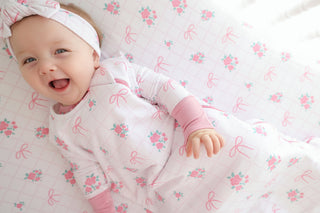 Infant sleep sack with white fabric featuring pink floral and bow print, pink ribbed trim, and matching headband.