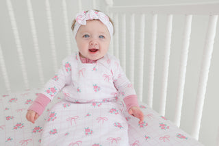 Infant in white sleep sack with pink floral and bow print, matching headband, lying in white crib.