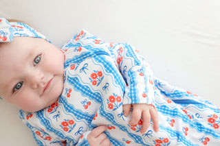 Infant in blue and red floral patterned sleep sack with bow and gingham details, wearing matching headband.
