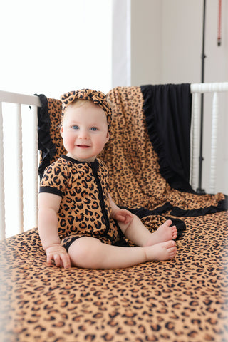 Baby sitting in crib wearing leopard print short-sleeve romper with matching headband and black trim.