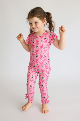 Child wearing pink short-sleeve pajama set with ribbon and figure print throughout, with ruffle details at ankles.