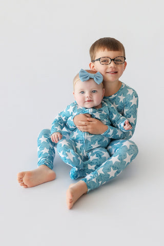 Infant wearing blue romper with white star pattern and matching bow headband, held by toddler in coordinating star-print paja