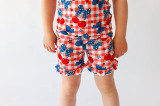 Child wearing red and white gingham shorts with cherry and bow print and ruffled leg trim.