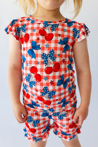 Child wearing red and white gingham short set with cherry and blue bow print, ruffled short sleeves and legs.