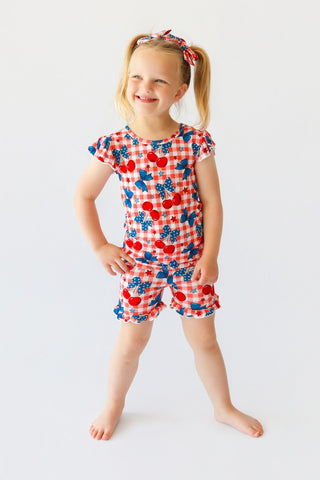 Girl wearing short-sleeved top and shorts with red cherry and blue star pattern on white gingham background.