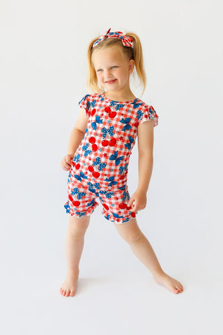 Girl wearing red and white checkered short set with cherry and bow print, ruffled sleeves and shorts, with matching hair bow.
