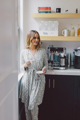 Woman wearing a striped robe with floral pattern in gray and pink tones, tied at waist, holding a mug in a kitchen setting.