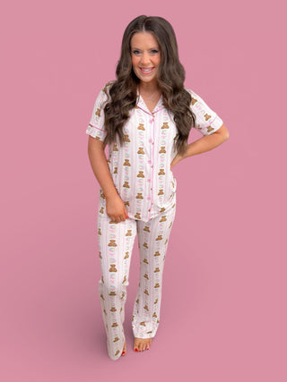 Woman wearing white short-sleeve pajama top and flare pants with brown bear and floral print throughout.