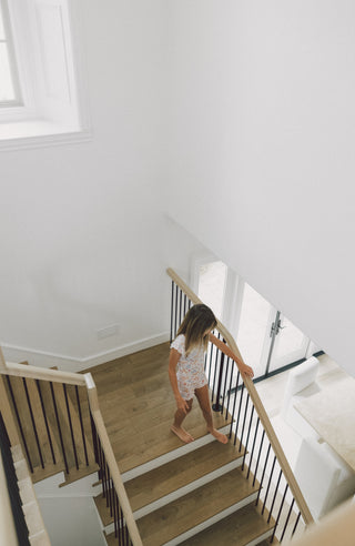 Child wearing white ruffle short set on wooden staircase with black metal railings.