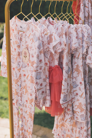Pink romper with horses, flowers, and boots print displayed on gold hanger with red ruffled detail.