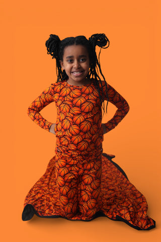 Child wearing orange basketball-print long-sleeve top and matching pants with black trim against orange background.