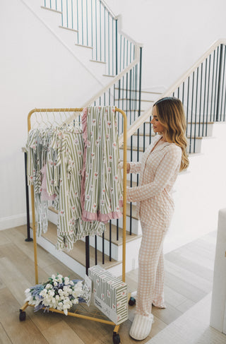Woman wearing beige and white gingham pajamas holding a clothing rack displaying striped and floral patterned pajama sets.