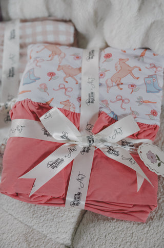 Pink ruffle blanket with boot and flower print, tied with white branded ribbon, displayed on light gray surface.