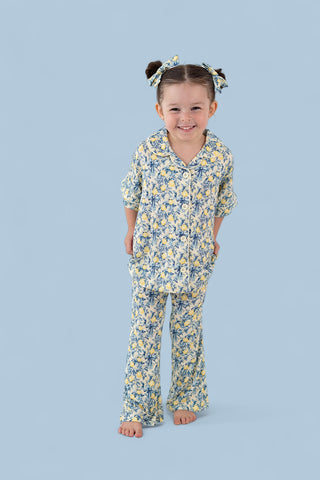 Young girl wearing yellow and blue citrus toile print button-up shirt and flared pants with matching hair bows.