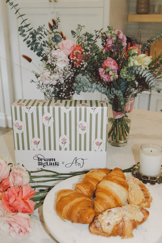 Striped gift box with floral pattern next to fresh flower arrangement and plate of croissants on white surface.