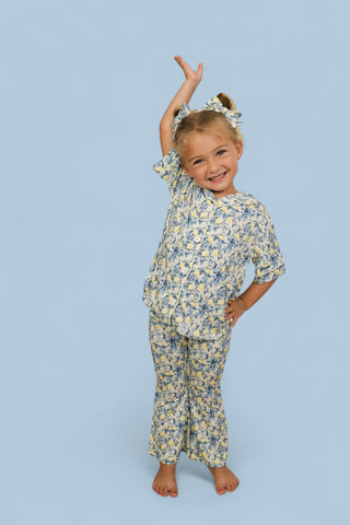 Young girl wearing yellow and blue citrus toile print short-sleeve top and flared pants with matching hair bow.