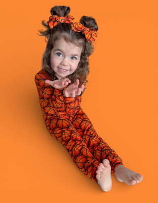 Young child wearing orange basketball-print pajamas with matching hair bows against orange background.