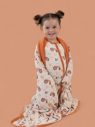 Young child wrapped in cream blanket with farmyard animal and tractor print, orange trim and zipper.