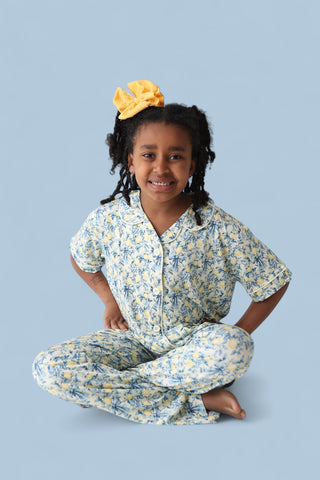 Child wearing yellow and blue citrus toile print short-sleeve button-up shirt and matching flare pants with yellow hair bow.
