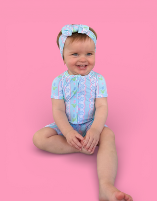 Toddler wearing light blue short-sleeve romper with tulip and geometric print, matching headband with bow.