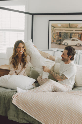 Woman in beige gingham pajama set and man in white shirt sitting on bed with pillows.
