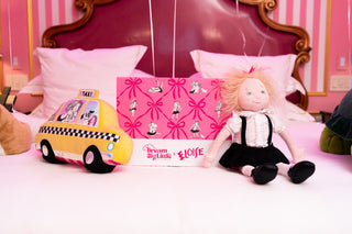Doll with blonde hair in black and white outfit next to yellow taxi toy and pink patterned gift box on bed.